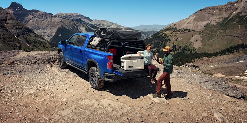 Wide rear side view of the 2025 Chevrolet Silverado on a mountain road. Two people are near the bed of the truck with a cooler.