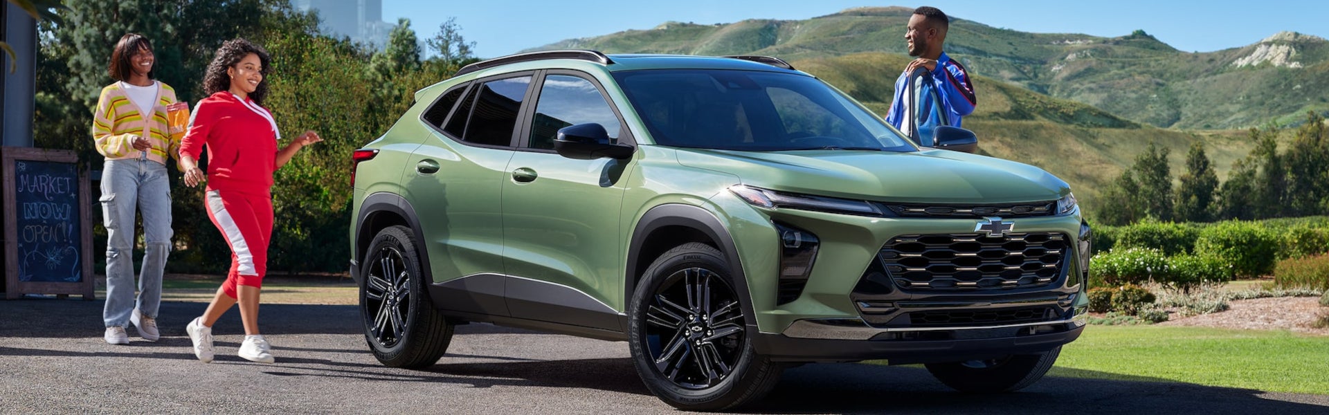 A family of three approaching a 2025 Chevy Trax which is parked in a sunny driveway with mountains in the background.