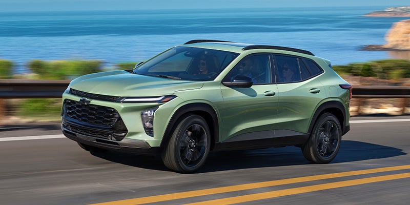 Side view of the 2025 Chevy Trax driving on a mountain road with a body of water in the background.